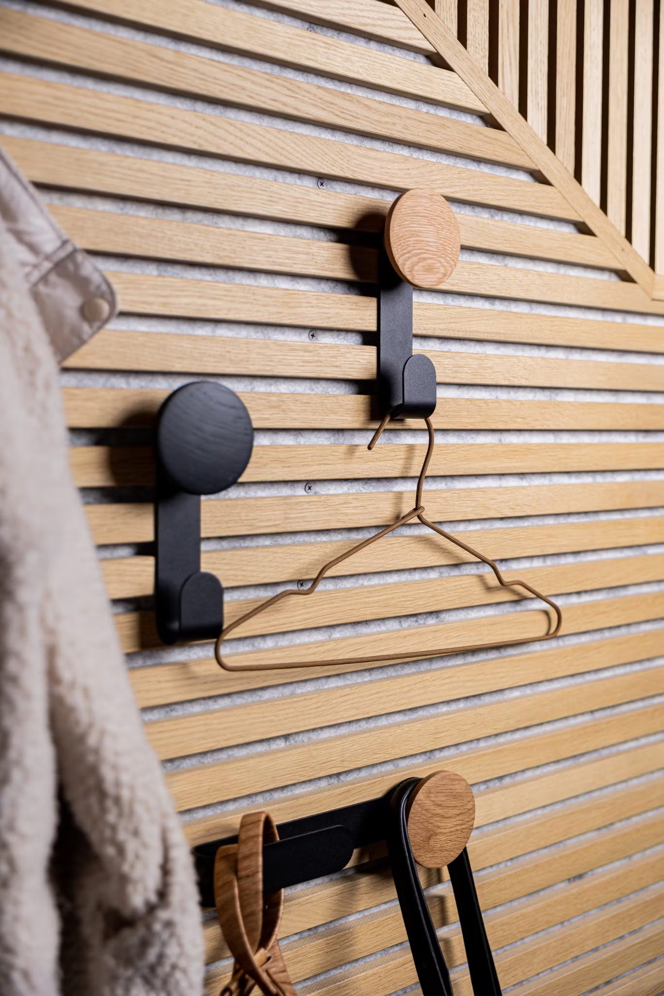 Tres modernos ganchos montados en una pared de listones de madera acústica Akupanel de estilo nórdico sujetan una percha metálica y accesorios, con un abrigo peludo parcialmente visible a la izquierda.